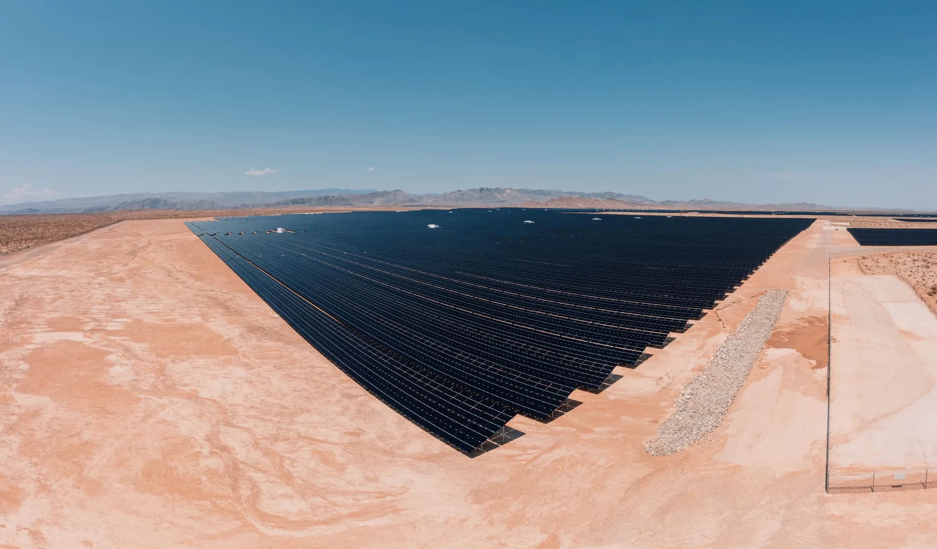 Aerial view of a solar farm built on brown desert sand, its rows of panels casting hard shadows under a clear sky.