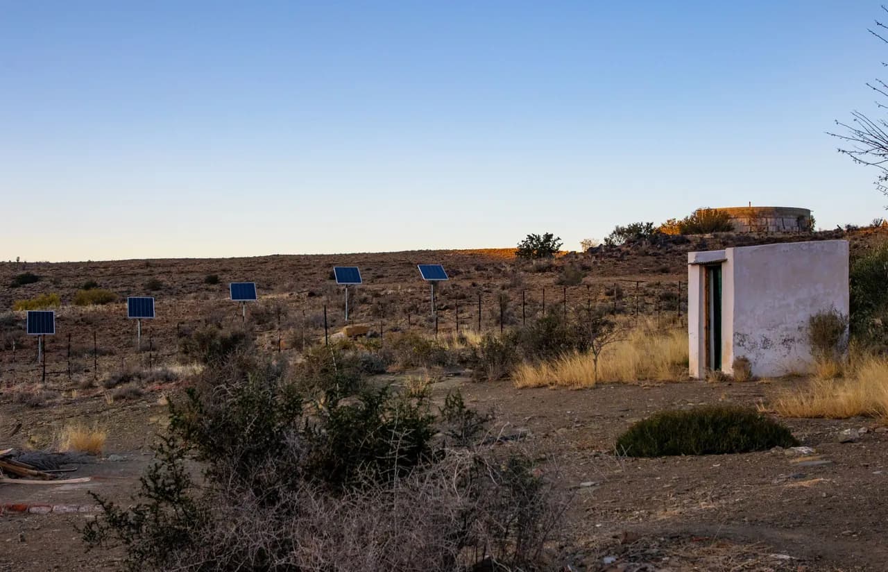 A small stand-alone solar array beside a lone shed in a dry desert landscape at sunset.
