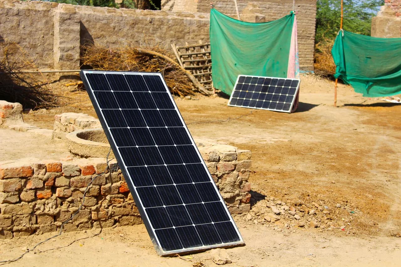 Two small solar panels propped on dry ground beside a circular brick-lined well in a rural yard — the minimal kind of array used to drive a water pump.